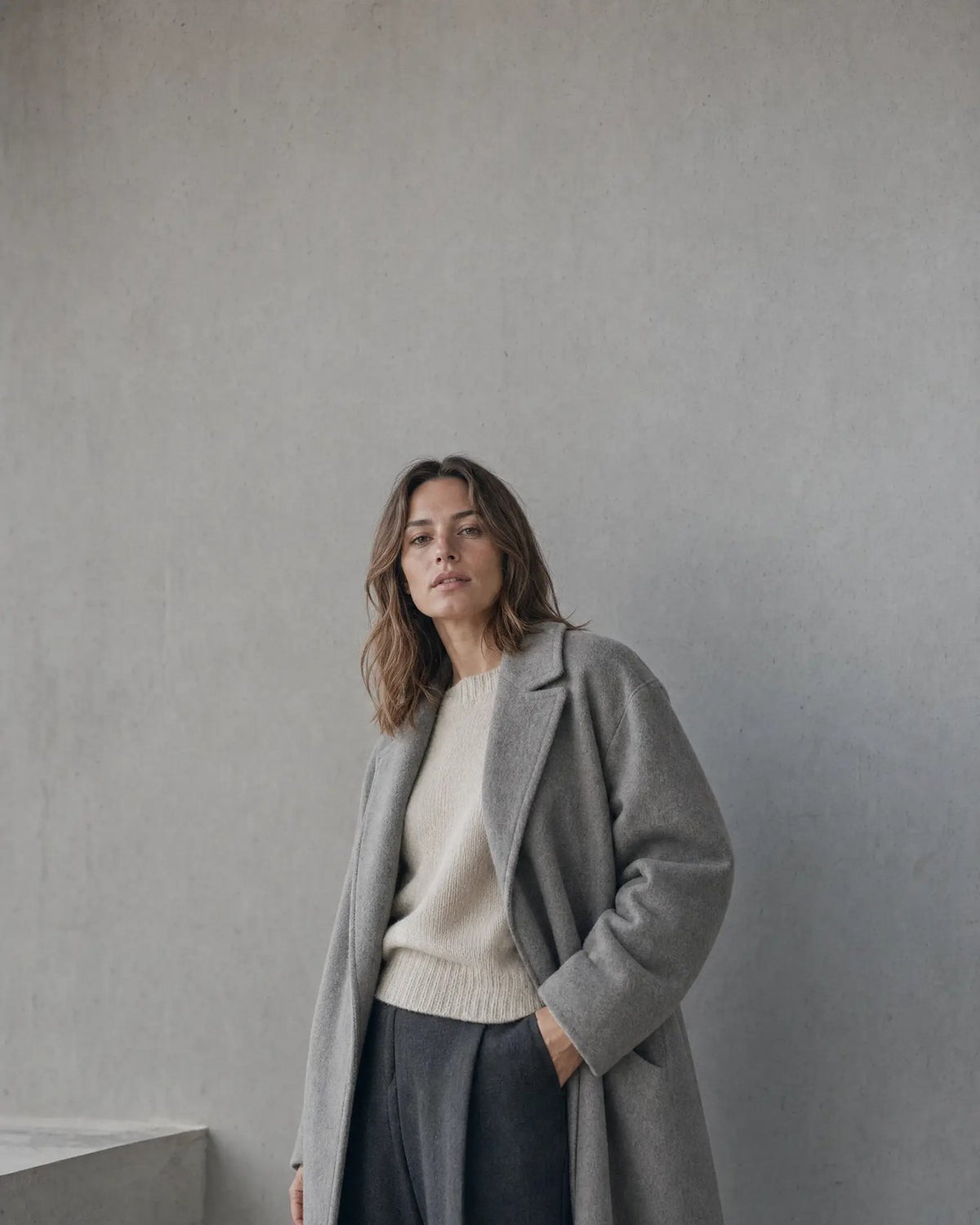 Frau in grauem Wollmantel über cremefarbenem Strickpullover vor heller Betonwand